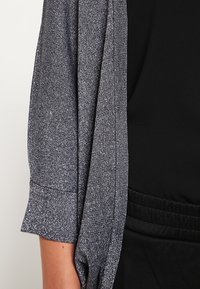 Stříbrný strukturovaný cardigan s krátkým rukávem, volným střihem a otevřeným předním dílem. V kombinaci s černým oděvem s elastickým páskem.