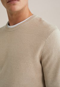 Bež pulover s teksturom i okruglim izrezom, s uzorkom saćaste mreže, od mekane tkanine, s bijelim kontrastnim unutarnjim ovratnikom.