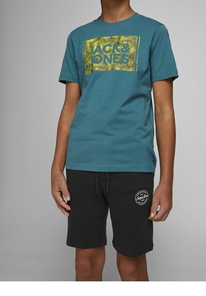 Garçon portant un t-shirt bleu canard Jack & Jones avec un logo jaune et un short noir avec un emblème circulaire blanc sur la jambe gauche, sur fond gris.