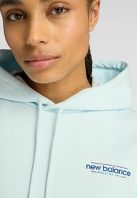 Sudadera azul claro hecha de material suave, con capucha ajustable y un logotipo en azul oscuro. Primer plano del escote y la cara.