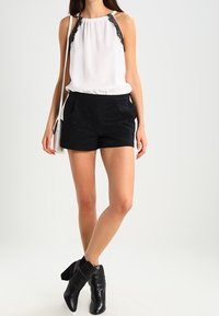 Blusa sin mangas blanca con detalles de encaje negro, combinada con pantalones cortos negros y botas de tobillo. Diseño simple, tela suave y pantalones cortos texturizados.