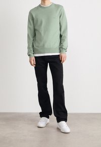 Grön stickad tröja med rundad hals, bärs över en vit skjorta. Svarta raka jeans och vita sneakers fulländar outfiten.