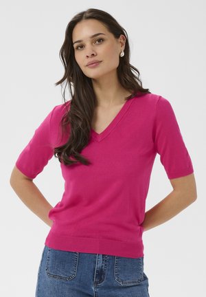 Kvinde med langt mørkt hår iført lyserød V-hals kortærmet sweater og blå jeans, stående med hænderne bag ryggen.