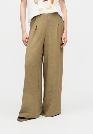 Φαρδιά, ψηλόμεση μπεζ παντελόνα με ρυτιδιασμένη υφή, συνδυασμένη με λευκό T-shirt με γραφικό σχέδιο και μαύρα παπούτσια με ταμπά σόλες.