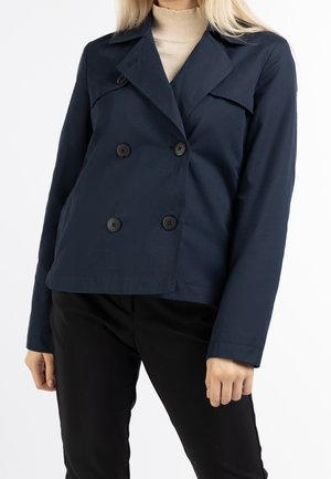 Kvinde iført en mørkeblå dobbeltradet blazer over en beige rullekrave og sorte bukser mod en ensfarvet hvid baggrund.