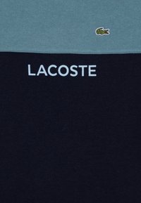 T-shirt à manches courtes, partie supérieure en bleu clair, partie inférieure en bleu marine, avec un petit logo de crocodile vert et le texte "LACOSTE" en blanc.