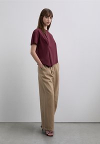 Kort shirt in bordeauxrood met korte mouwen, een losse pasvorm en een schuin inzetten detail, gecombineerd met beige wijduitlopende broek en bordeauxrode sandalen met bandjes.