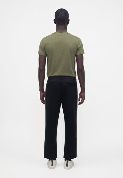Homme debout, de dos, portant une chemise à manches courtes vert olive, un pantalon noir avec une poche arrière et des baskets blanches avec des rayures noires et vertes.
