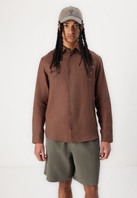 Camisa marrón de manga larga con botones en el frente, combinada con pantalones cortos a rayas verdes. El modelo lleva una gorra de béisbol gris. Atuendo sencillo y casual.