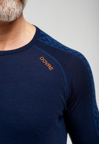 Marineblå langærmet skjorte med mønstrede blå detaljer på skuldrene. Har orange "DOVRE" branding på brystet. Glat, tætsiddende tekstur.