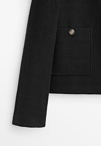 Svart texturerad jacka med en enkel framficka med en dekorativ knapp och en rak ärmdesign.