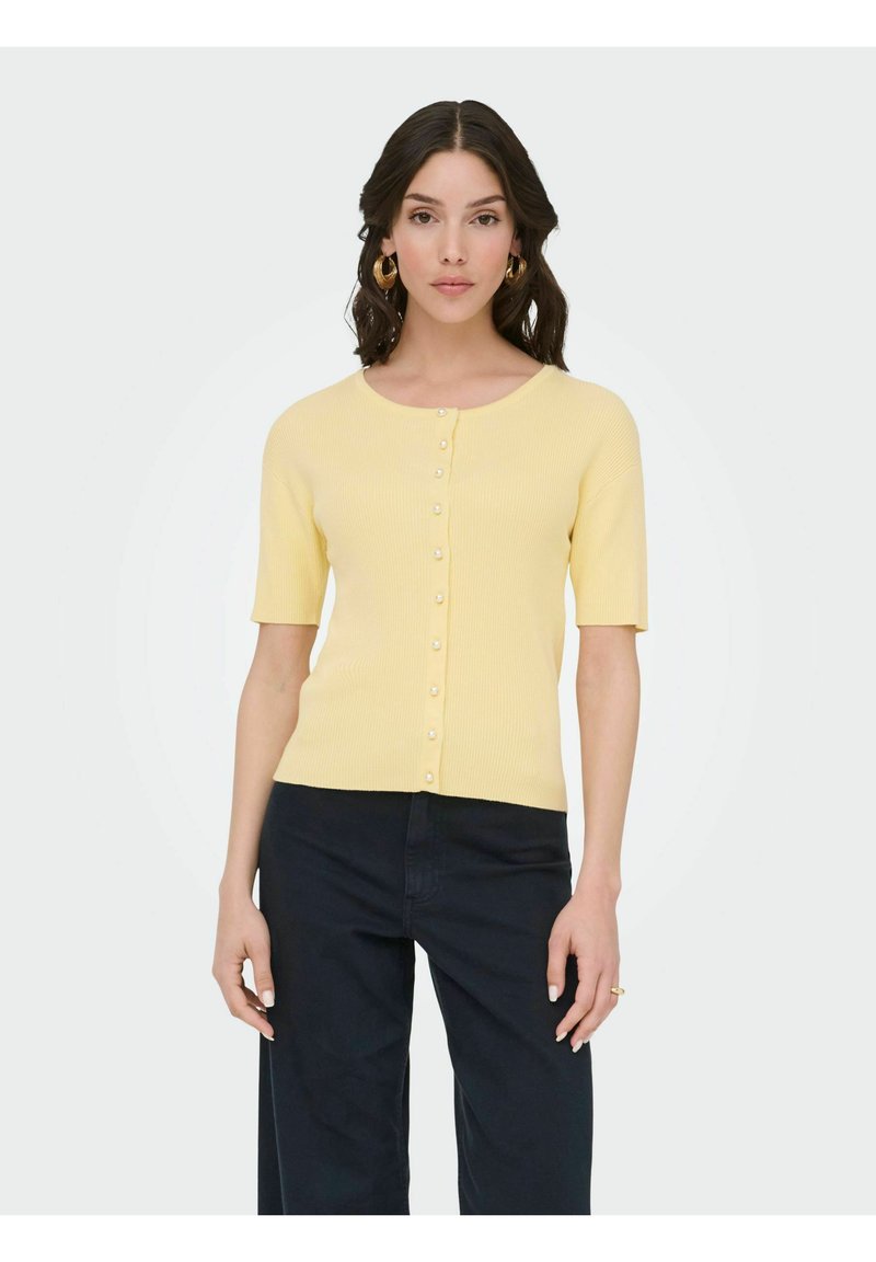 Femme portant un cardigan à manches courtes jaune pâle boutonné et un pantalon large sombre, debout devant un fond clair uni.