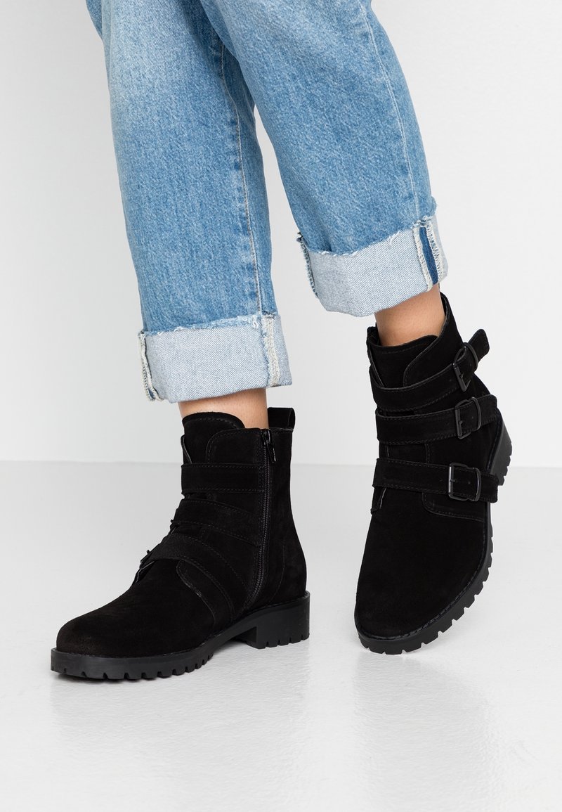 Botines de ante negros con tres correas ajustables, un zipper lateral y una suela de goma gruesa. Llevados con vaqueros azules doblados.