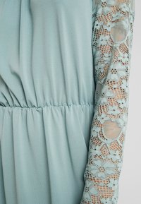 Robe bleu clair avec un corsage texturé et des manches longues en dentelle complexe ornée de motifs floraux, détail de taille froncée.