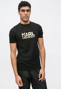 Camiseta negra de manga corta, con un prominente logo dorado "KARL LAGERFELD" en tipo de letra negrita, tela suave y cuello redondo.