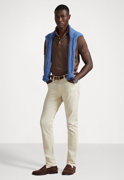 Conjunto masculino com uma camisola polo castanha, calças em bege claro, um suéter azul de malha trançada pousado nos ombros, e mocassins em camurça castanha.