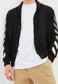 Cardigan nero a maglia con colletto a scialle, dotato di sei bottoni neri, polsini a coste e motivi a zigzag bianchi e grigi sulle maniche.