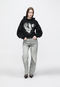 Black hoodie with a graphic design of a skull and leaves. Paired with light grey wide-leg jeans and black pointed-toe heels.