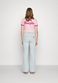 Kobieta stojąca tyłem, ubrana w jasnoniebieskie dżinsy z szerokimi nogawkami i różową koszulkę z czerwoną napisem "Wrangler" na plecach.