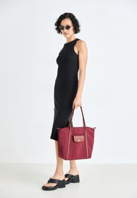 Maroon tote bag with a brown accent, made of fabric with handles. Worn with a fitted black dress and platform sandals, against a light background.