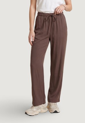 Persona che indossa pantaloni marroni larghi con coulisse, scarpe da ginnastica bianche e una maglietta bianca, in piedi davanti a uno sfondo chiaro e uniforme.