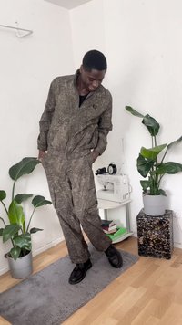 Macacão com padrão de camuflagem em tons terrosos suaves, com fecho na frente, corte solto e bolsos laterais, combinado com sapatos pretos com atacadores.