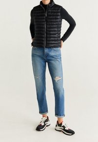 Gilet matelassé noir sur une chemise noire à manches longues, associé à un jean bleu déchiré et des baskets noires avec des accents rouges.