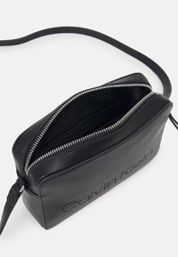 Sort skuldertaske i læder med lynlåslukning, der har en glat tekstur og et diskret præget logo. Inkluderer en tynd, justerbar rem.