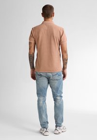 Polo tričko v světle broskvové barvě s texturovaným vzorem, spárované s světle modrými džíny. Boty jsou bílé s námořnickými akcenty. Pohled zezadu.