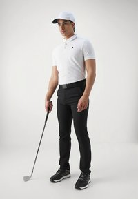 Hvid polo med sort logo, sorte skræddersyede bukser og sorte golfsko. Figuren holder en golfkølle og bærer en hvid kasket.