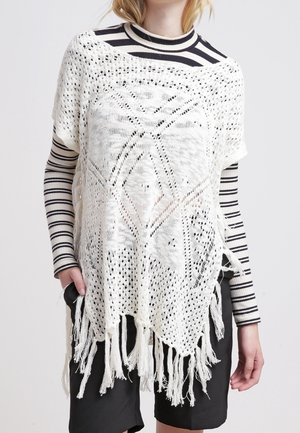Femme portant un poncho blanc crocheté avec des franges par-dessus un haut à manches longues rayé noir et blanc et un short noir.