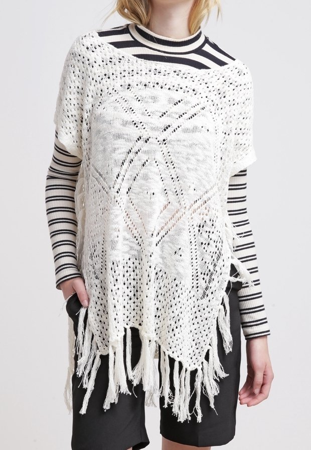Femme portant un poncho blanc crocheté avec des franges par-dessus un haut à manches longues rayé noir et blanc et un short noir.