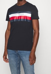 Černé bavlněné tričko s horizontálním pruhovaným grafickým vzorem v červené, bílé a námořnické modré barvě, se znakem "TOMMY HILFIGER" na přední straně.