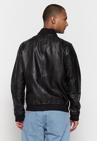 Uomo che indossa una giacca di pelle nera e jeans azzurri chiari, di spalle con la testa girata a destra su uno sfondo bianco uniforme.