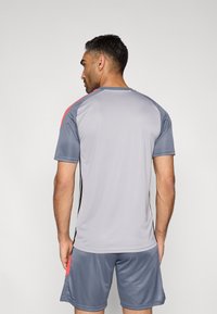 Γκρι αθλητική μπλούζα με κοντά μανίκια και στρογγυλή λαιμόκοψη, με μαύρες ρίγες στα πλάγια και κόκκινες λεπτομέρειες στους ώμους, συνδυασμένη με γκρι σορτς.