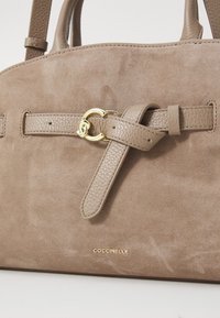 Sac à main en suède beige clair avec des accents en cuir texturé, une bandoulière à l'avant soigneusement nouée et un fermoir circulaire en ton doré. Logo embossé à l'avant.