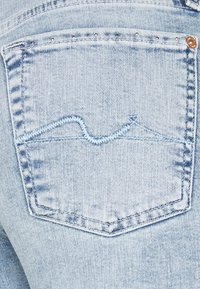 Bolsillo de jeans de mezclilla azul claro con diseño bordado ondulado en un hilo en contraste. Presenta remaches de metal a lo largo del borde del bolsillo.