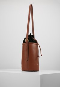 Borsa a secchiello in pelle marrone con tracolla regolabile e chiusura a cordoncino nera, in piedi su un piedistallo bianco.