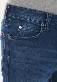 Los jeans de mezclilla azul oscuro presentan un diseño de cinco bolsillos con herrajes metálicos, costuras en contraste y una textura suave.