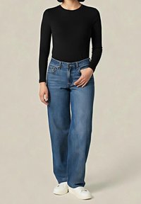 Personne portant un haut noir à manches longues, un jean bleu taille haute droit et des chaussures blanches sans lacets, debout devant un fond uni.