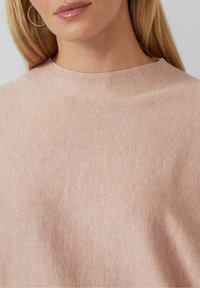 Gros plan sur une femme portant un pull en maille rose pâle et une boucle d'oreille argentée en forme d'anneau, avec de longs cheveux blonds visibles.