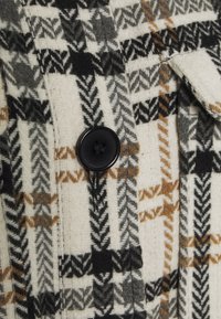 Tessuto misto lana con base color crema e motivo pied de poule in nero, grigio e marrone. Caratterizzato da un grande bottone nero e dettaglio con tasca.