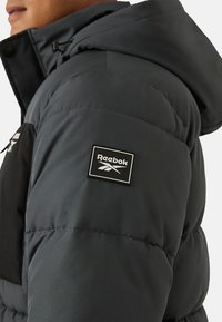 Zwart en donkergrijs donsjack met capuchon, voorzien van een Reebok-logopatch op de schouder en een doorgestikte textuur.