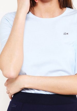 Vrouw die een lichtblauw shirt met korte mouwen draagt met een klein geborduurd krokodillenlogo op de bovenste linker borst, gecombineerd met donkere broek.