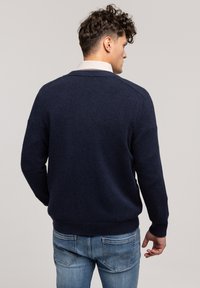 Granatowy sweter dzianinowy z prążkowanymi mankietami i dołem, z okrągłym dekoltem. Noszony na koszuli z kołnierzykiem, pokazany z tyłu.