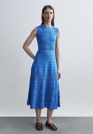 Žena stojí oblečená v modrých bezrukávových midi šatách s textúrou, ktoré sú spojené s leopardími plochými topánkami, na jednoduchom sivom pozadí.