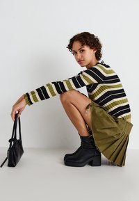 Pull en tricot rayé vert, noir et crème ; jupe olive plissée ; bottes à plateforme noires à la cheville ; tenant un petit sac à main noir.