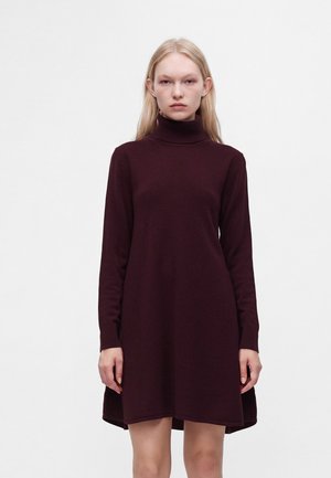 Jeune femme aux cheveux blonds lisses portant une robe à col roulé bordeaux foncé à manches longues sur un fond blanc uni.