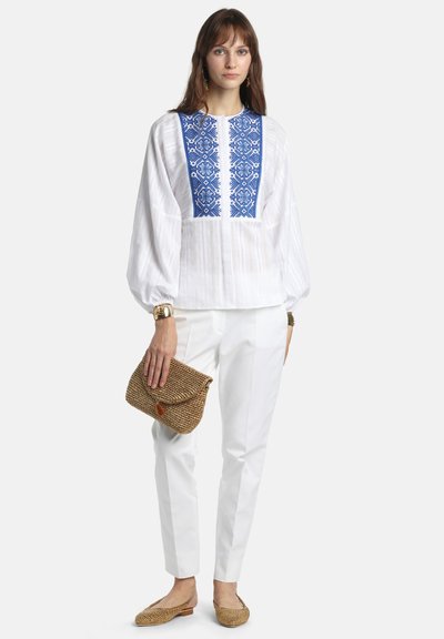 Femme portant un chemisier blanc avec un panneau avant bleu brodé, un pantalon blanc, tenant une pochette tissée et des chaussures plates tissées assorties, debout face à l'avant.