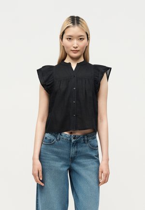 Jeune femme aux cheveux blonds et lisses porte un top noir sans manches boutonné avec des volants aux épaules et un jean bleu taille haute.